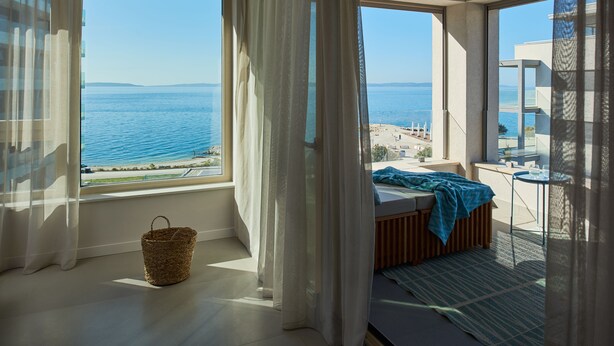 A bedroom with a large window overlooking the sea, a bed, a small table, and a basket on the floor. - Eol Apartments Split (Split)