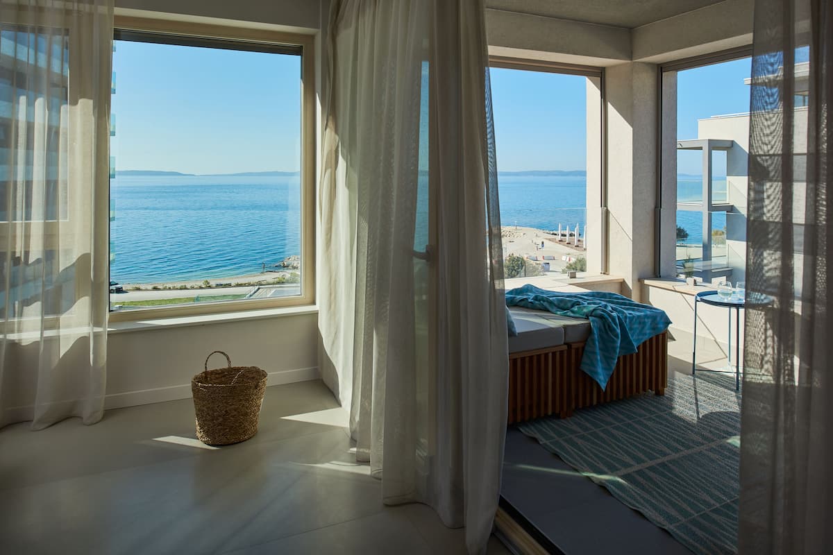 A bedroom with a large window overlooking the sea, a bed, a small table, and a basket on the floor.