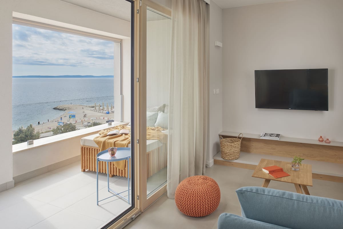 A modern living room with a large window overlooking a beach, a flat-screen TV mounted on the wall, and a cozy seating area with a blue sofa and a round orange ottoman.