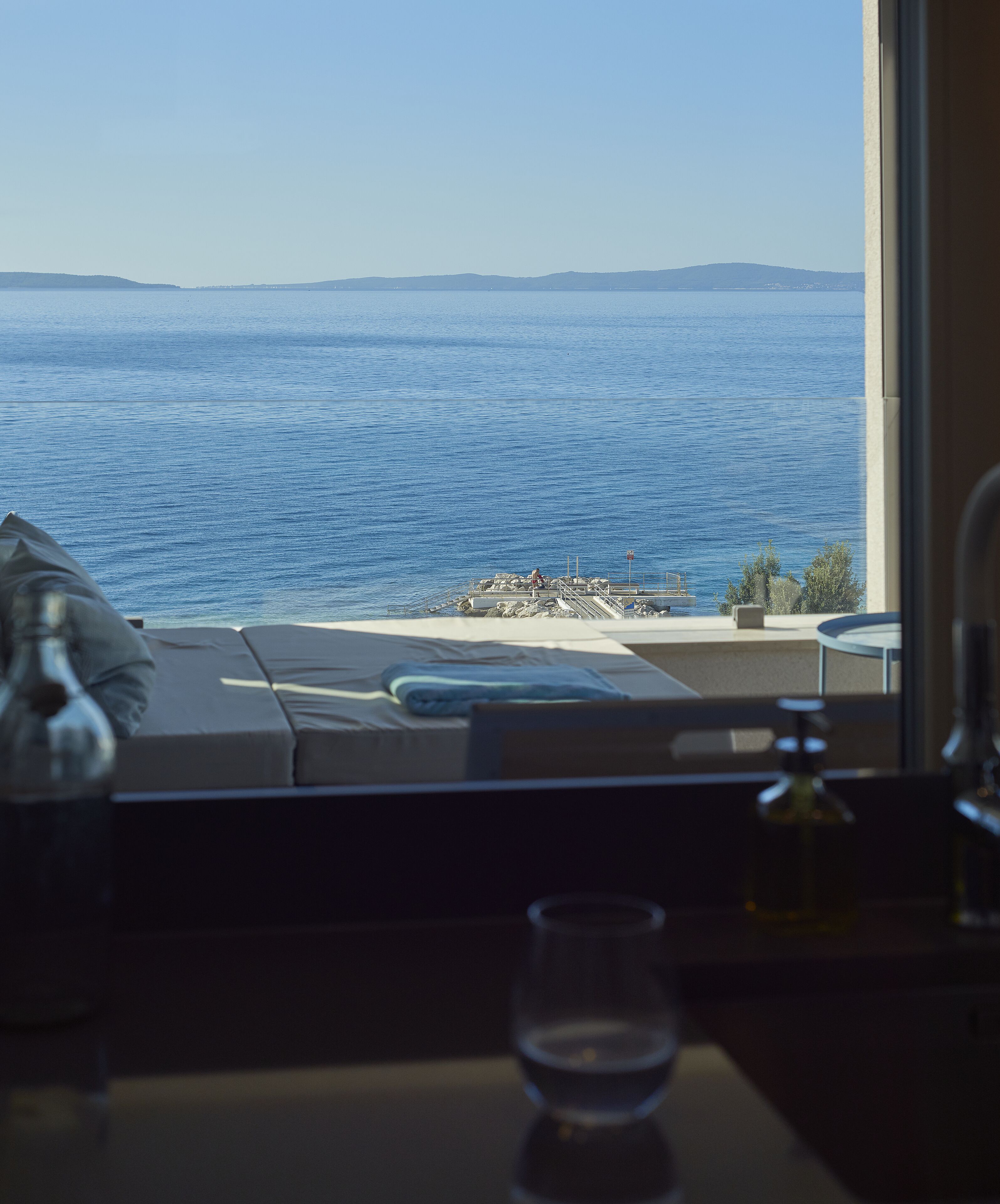 A room with a view of the sea, a sofa, a table with a glass, and a bottle.