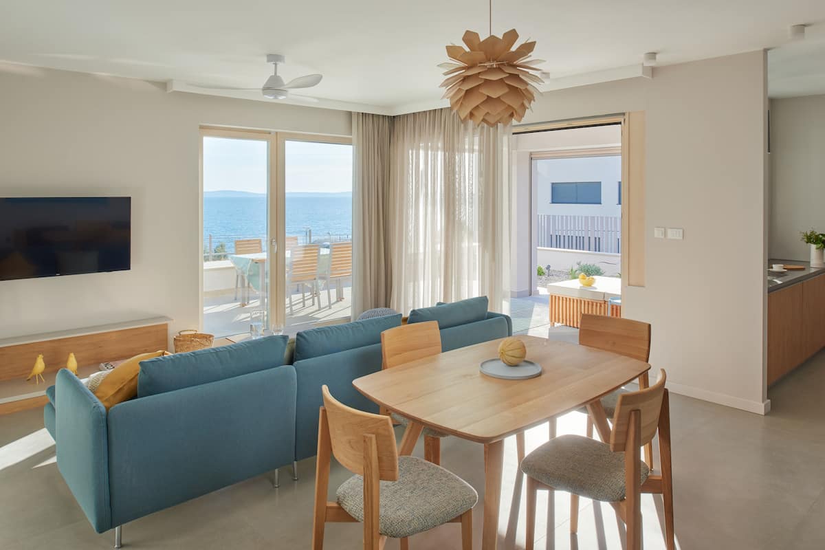 A modern living room with a blue sofa, wooden dining table, and chairs, a flat-screen TV, and a view of the sea through large windows.