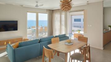 A modern living room with a blue sofa, wooden dining table, and chairs, a flat-screen TV, and a view of the sea through large windows.