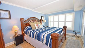 A bedroom with a large bed, a nightstand with a lamp, a window with blinds, and a framed picture on the wall. - Ocean Hideaway 404-Oceanfront 18th St, Free WiFi, Elev, W/D, AC (Ocean City)