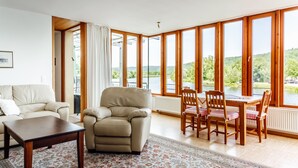 A spacious living room with a sofa, armchair, and dining table with chairs. Large windows offer a view of a lake and greenery. - FeWo Schloß Ramspau - Forsthaus Ostwohnung (5-star DTV) (Ramspau)