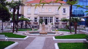 Un edificio con una fuente en el centro, rodeado de un jardín bien cuidado con palmeras y un sendero pavimentado.