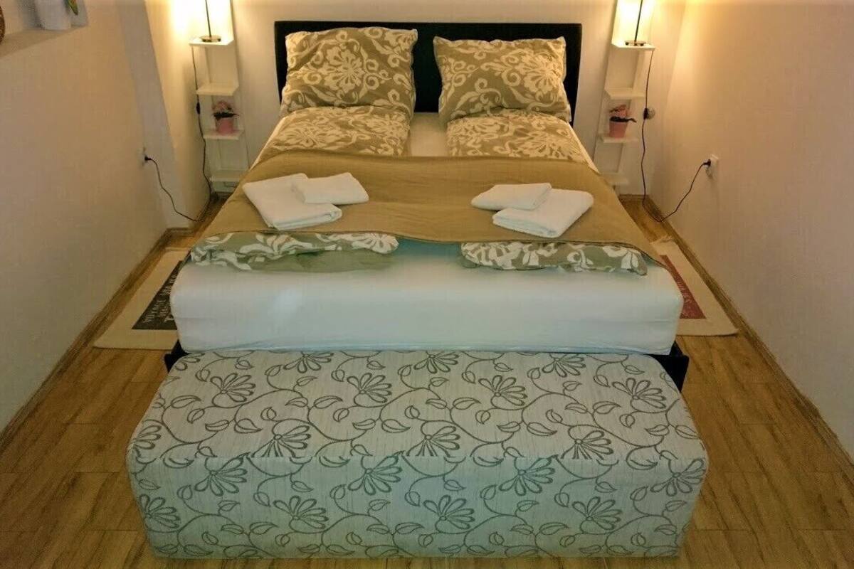A neatly made bed with floral-patterned bedding and pillows, a bedside table with a lamp, and a small shelf with decorative items.