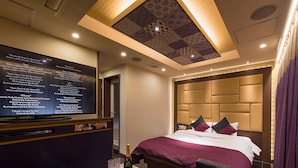 Una habitación de hotel moderna con una amplia televisión de pantalla plana mostrando una lista de canciones, una cama cómoda con cabecera y un techo con un diseño geométrico.