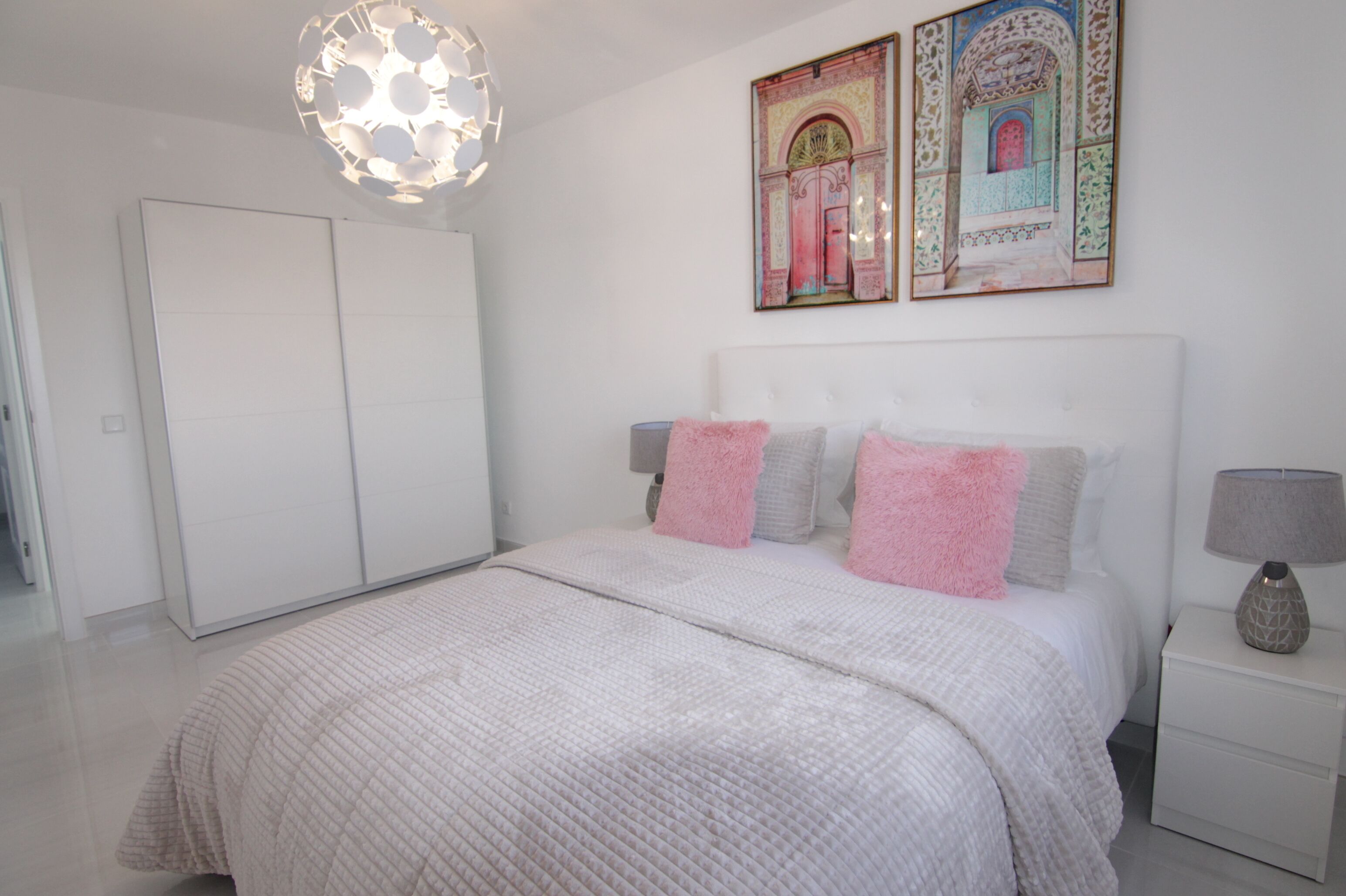 Een slaapkamer met een groot bed, een kledingkast, een nachtkastje met een lamp en twee aan de muur hangende schilderijen.