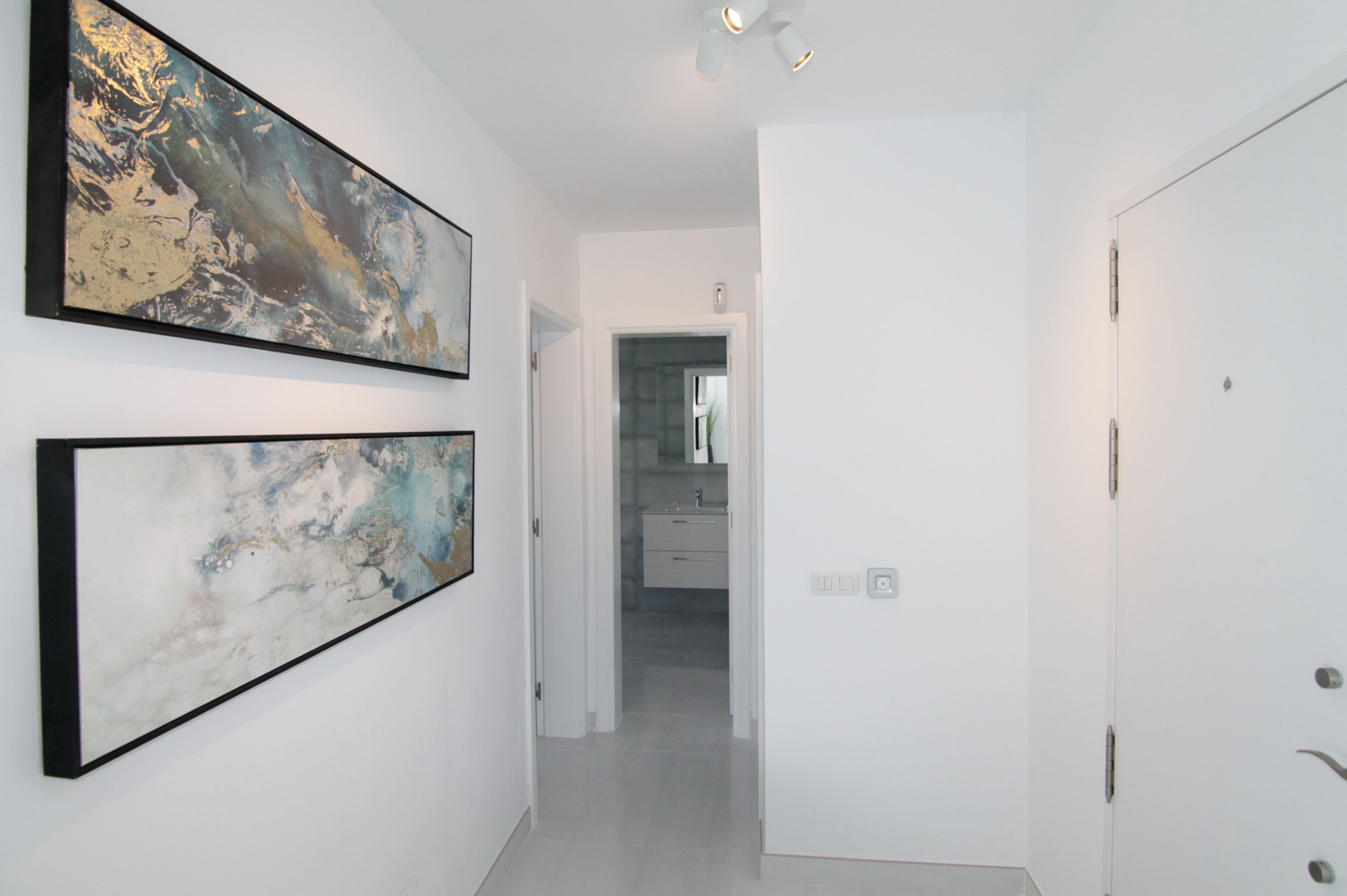 A hallway with two abstract paintings on the walls, leading to a bathroom with a sink and mirror.