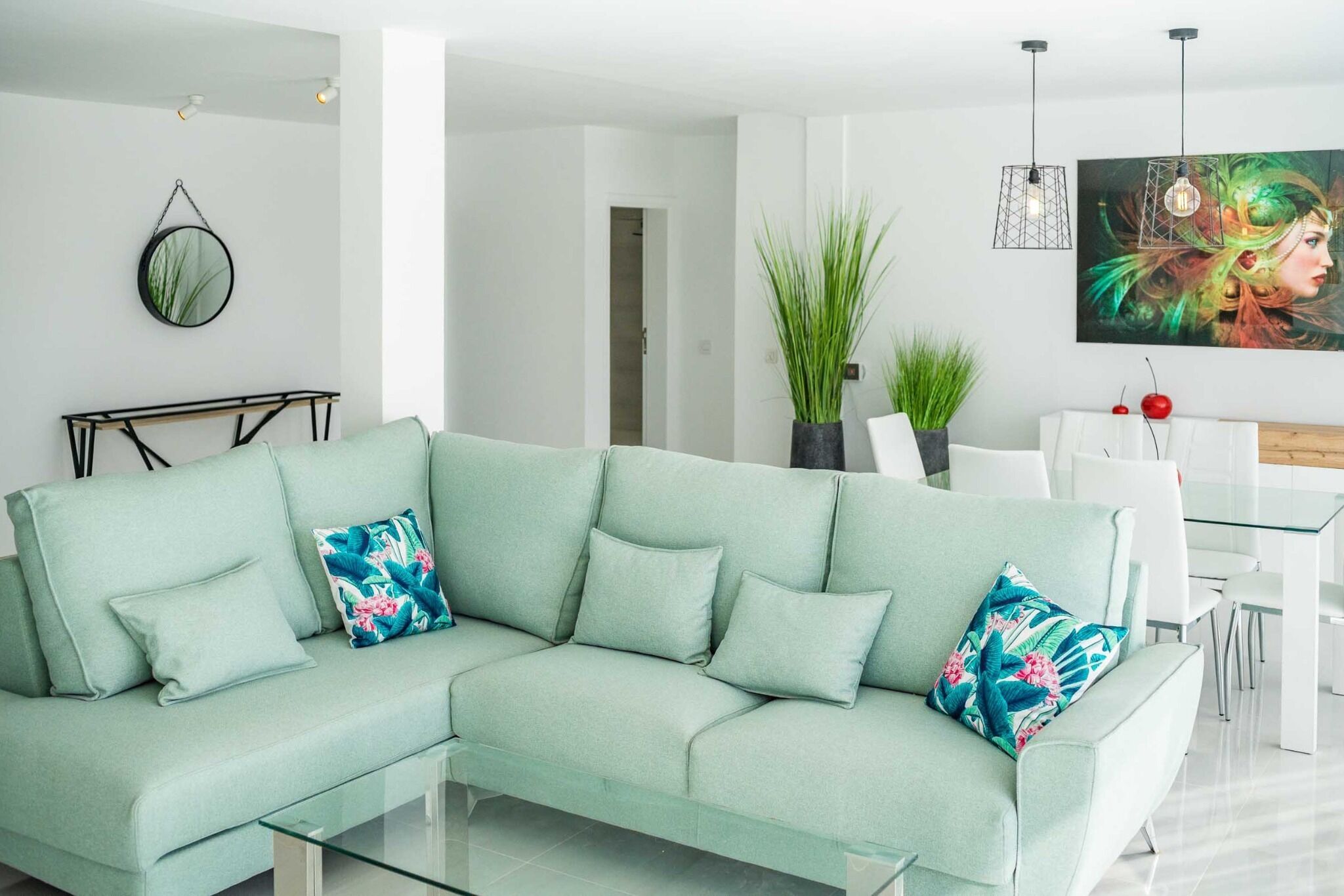 A modern living room with a light green sectional sofa, a glass coffee table, and a dining area with white chairs and a glass table.