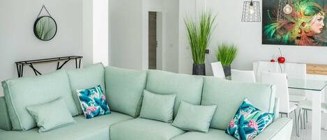 A modern living room with a light green sectional sofa, a glass coffee table, and a dining area with white chairs and a glass table.
