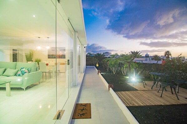 Un intérieur moderne avec un mur de verre, un canapé vert et une terrasse en bois aménagée avec du mobilier d’extérieur.