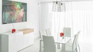 Een moderne eethoek met een glazen tafel, witte stoelen, een wit kastje en een hanglamp.
