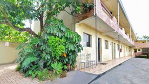 A narrow alleyway with a building on one side and a tree on the other. There are two white chairs outside the building. - Pousada Aconchego (Peruíbe)