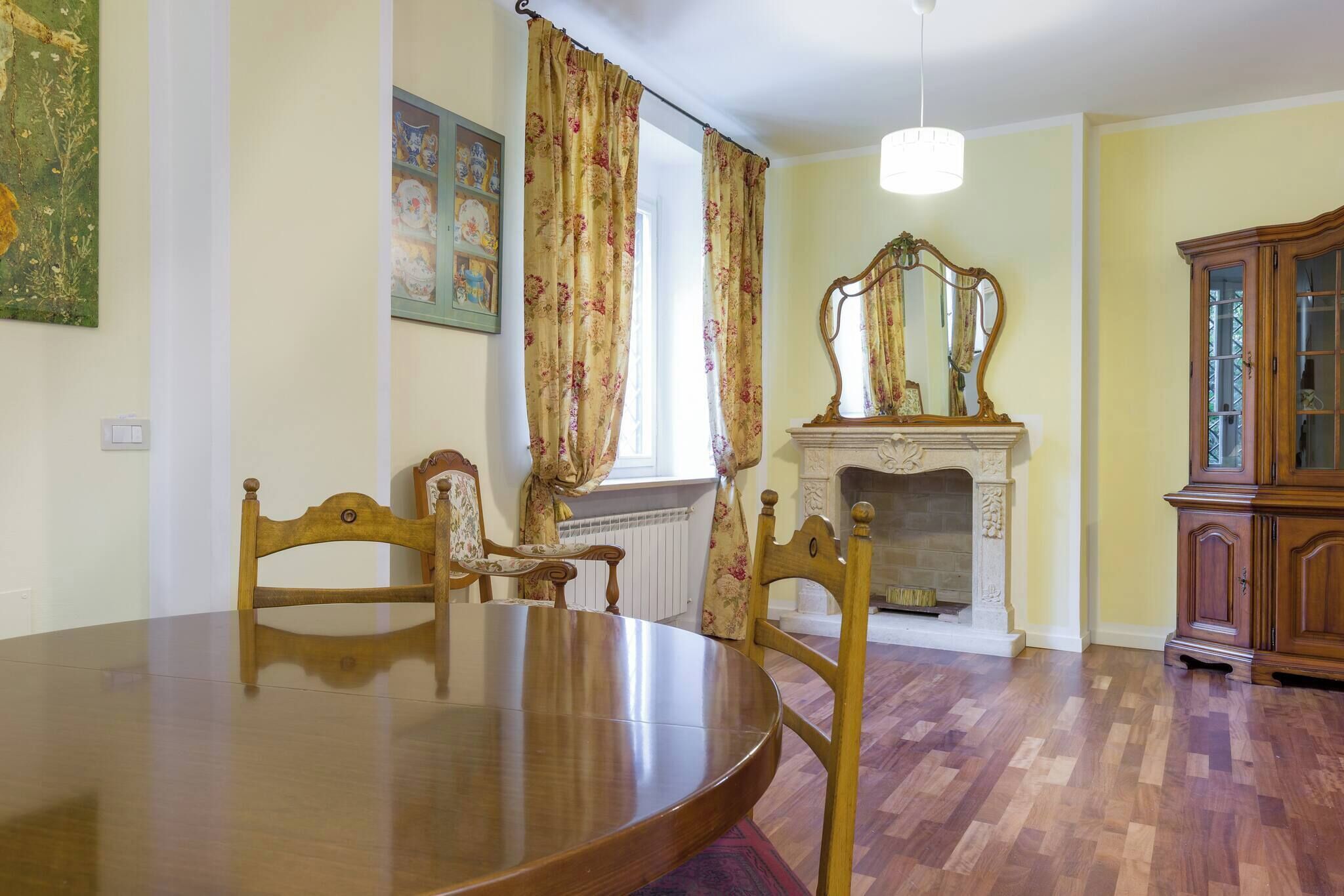 A dining area with a wooden table and chairs, a fireplace with a mirror above it, and a cabinet to the right.