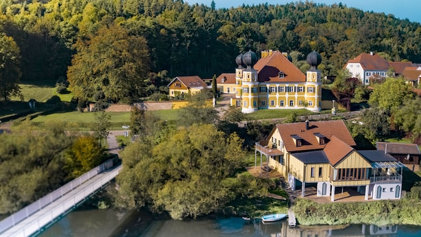 A large, yellow building with multiple towers, surrounded by lush greenery and a river.