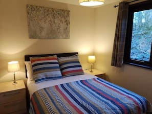 A neatly made bed with striped bedding, two bedside lamps, a nightstand with a drawer, and a window with curtains.