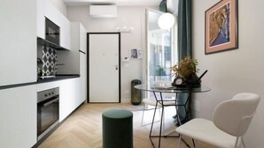 Ett kompakt kök med vita skåp, en svart bänkskiva och en modern spis. Här finns en matplats med ett glasbord och vita stolar, samt ett stort fönster med utsikt över staden.