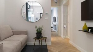 Ett vardagsrum med en beige soffa, en rund spegel, ett litet bord med en växt och en tv på väggen.