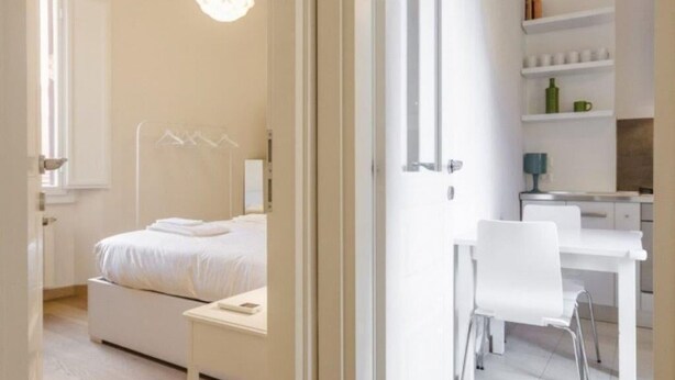 Una habitación de hotel moderna con cama, mesita de noche y escritorio con silla. Se observa una estantería con libros y una botella verde.