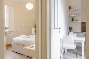 A modern hotel room with a bed, bedside table, and a desk with a chair. There is a shelf with books and a green bottle.