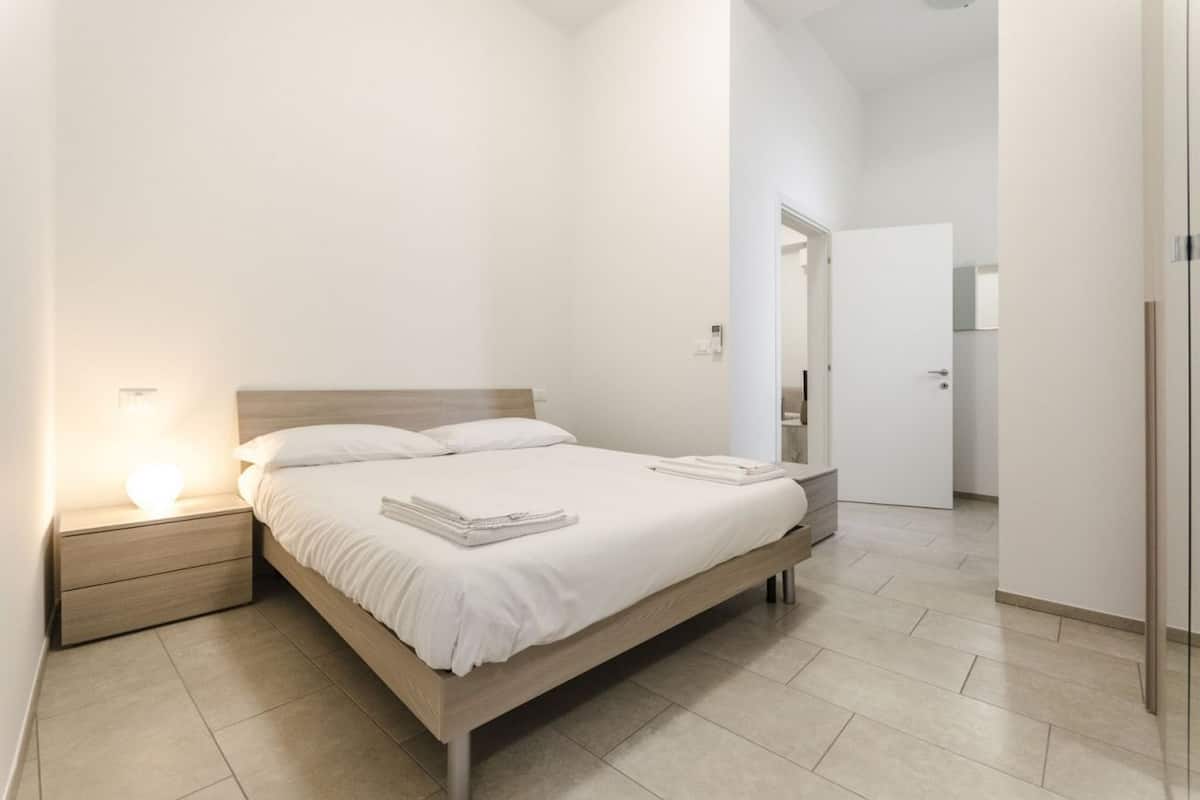 A modern bedroom with a wooden bed, a bedside table with a lamp, and a door leading to another room.