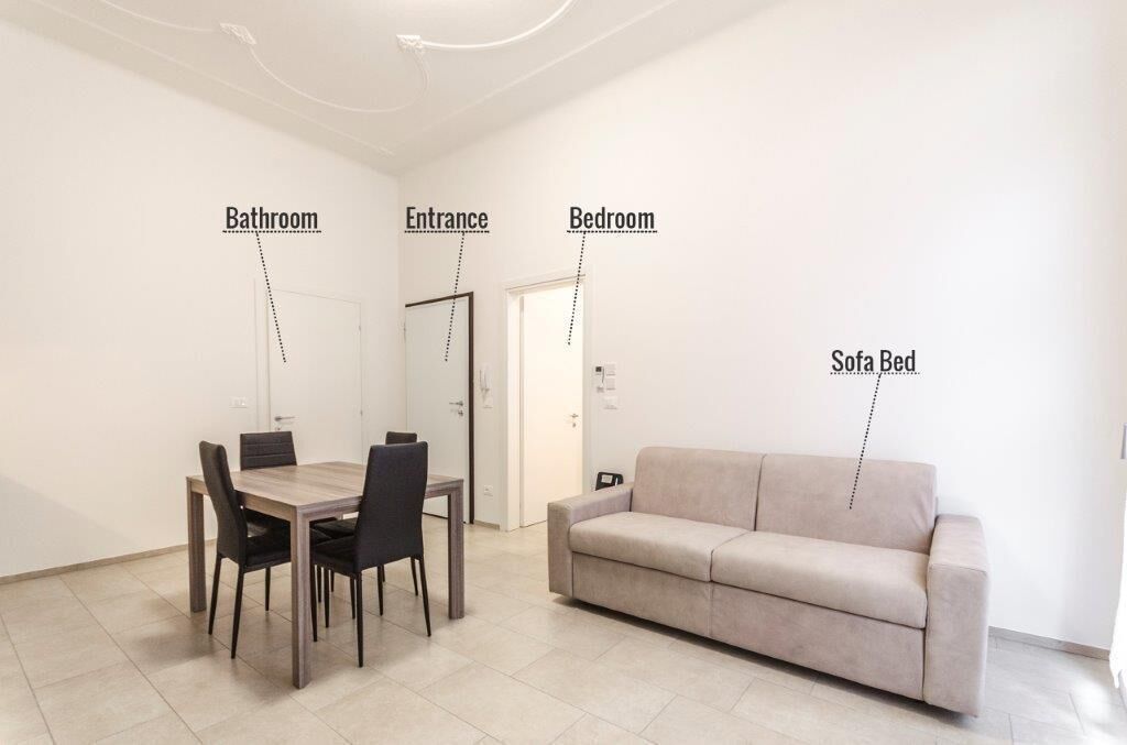 Habitación minimalista con una mesa de comedor y sillas, una cama sofá y zonas delimitadas para el baño, la entrada y el dormitorio.