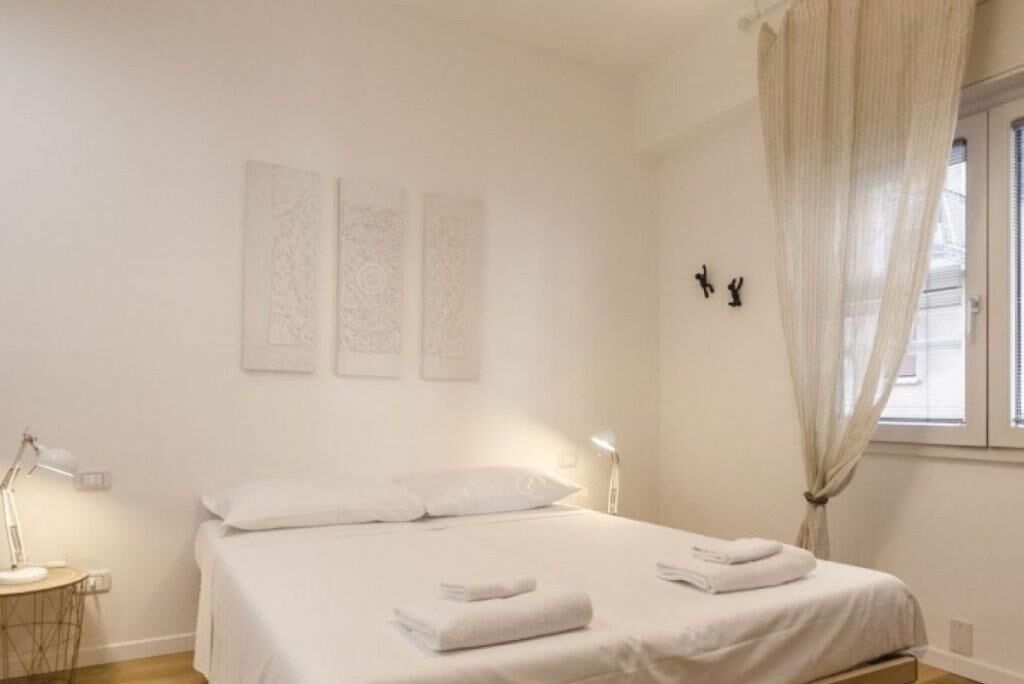 A clean, minimalist bedroom with a bed, bedside lamps, and a window with sheer curtains.