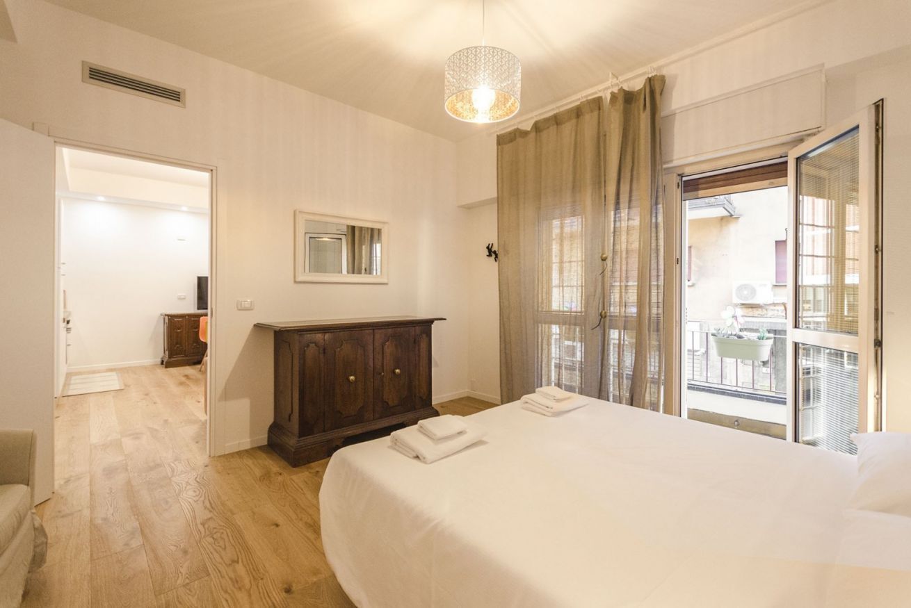 Quarto com cama grande, penteadeira de madeira, janela com cortinas e vista para varanda externa.