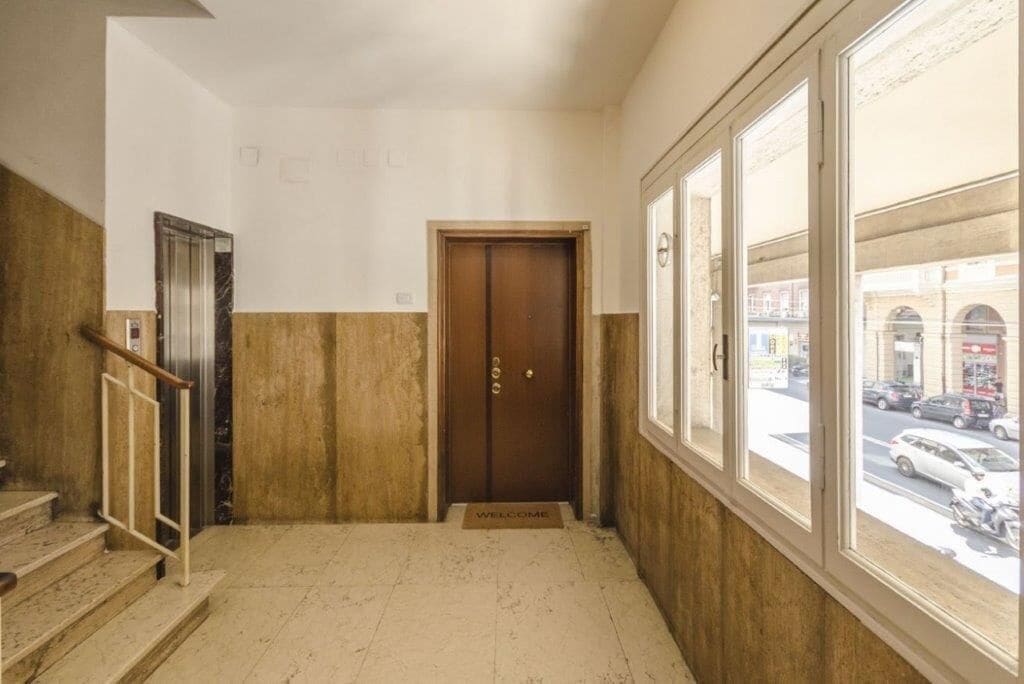 Un pasillo con una puerta de madera, un ascensor metálico y una ventana con vista a la calle.