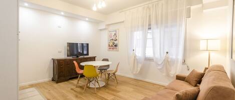 A living room with a brown sofa, a dining table with chairs, a TV, and a poster on the wall.