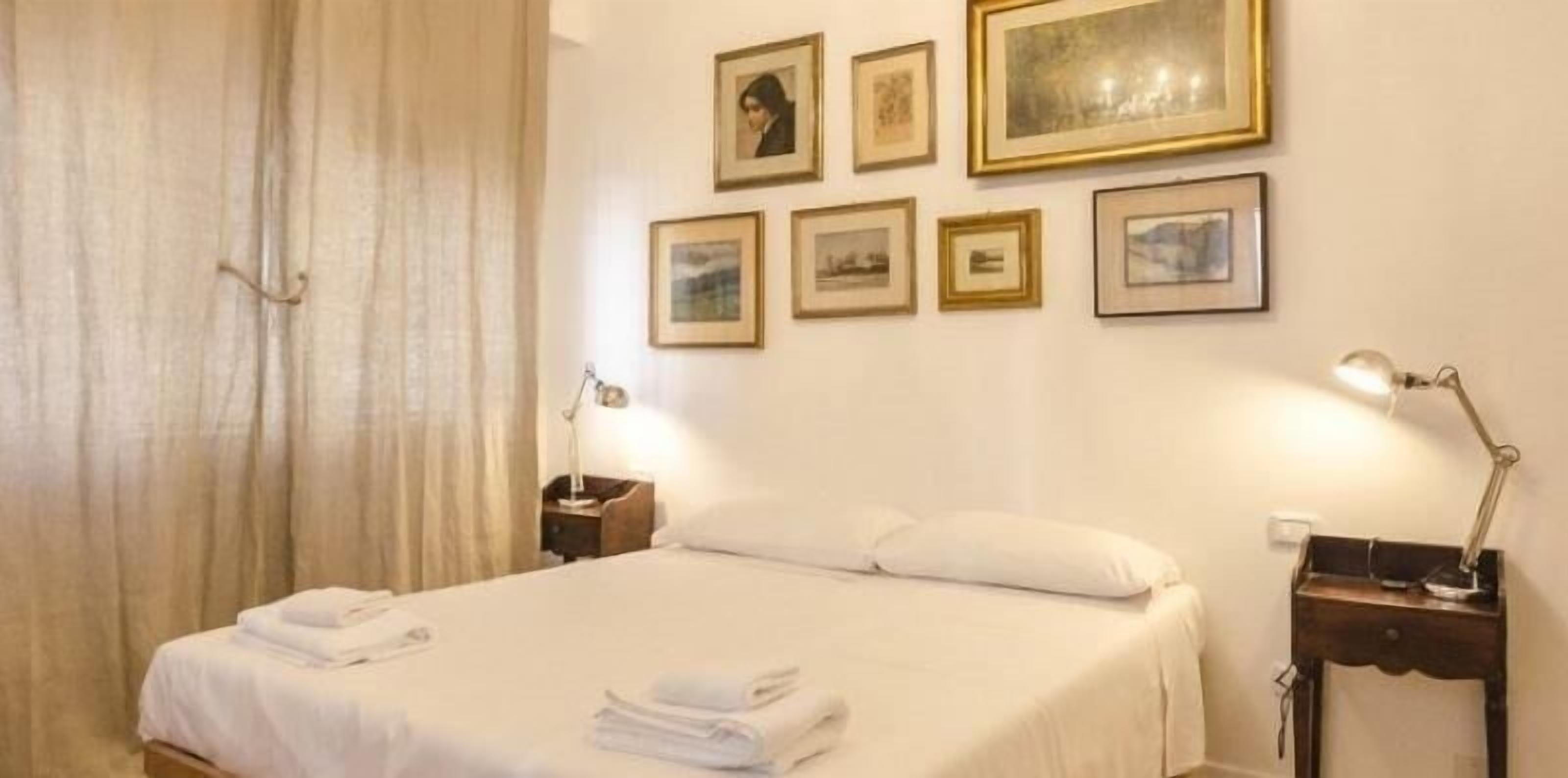 Una habitación de hotel con una cama, dos mesitas de noche, una lámpara y varias fotografías enmarcadas en la pared.