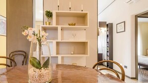 Un comedor con una mesa redonda, sillas, un florero con flores y una estantería blanca decorada con objetos.