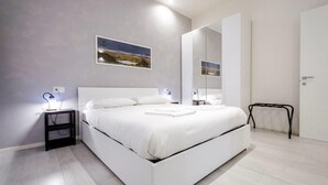 Ein modernes Schlafzimmer mit einem weißen Bett, einem Nachttisch mit Lampe, einem großen Spiegel und einem Gemälde an der Wand.