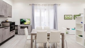 Eine moderne Küche mit grauen Schränken, einem Esstisch mit vier Stühlen, einem Fernseher auf einem Lowboard und einem Fenster mit Vorhängen.