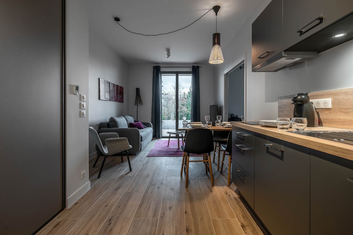 Une cuisine moderne avec des meubles en bois, un coin repas comprenant une table et des chaises, et une vue sur l’extérieur grâce à une grande fenêtre.