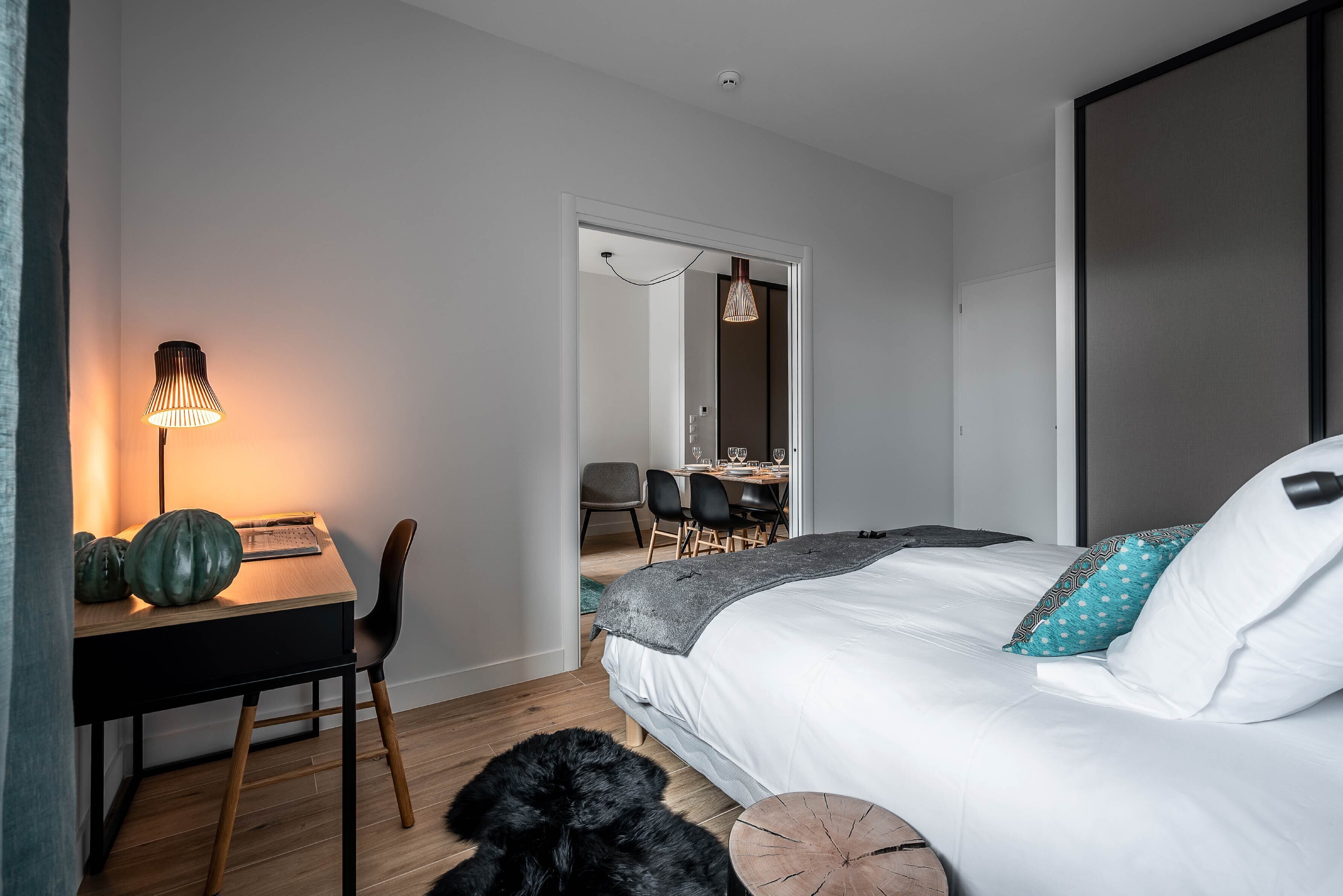 Camera da letto moderna con un letto, una scrivania e una sedia. Si intravede un'area pranzo attraverso una porta aperta.