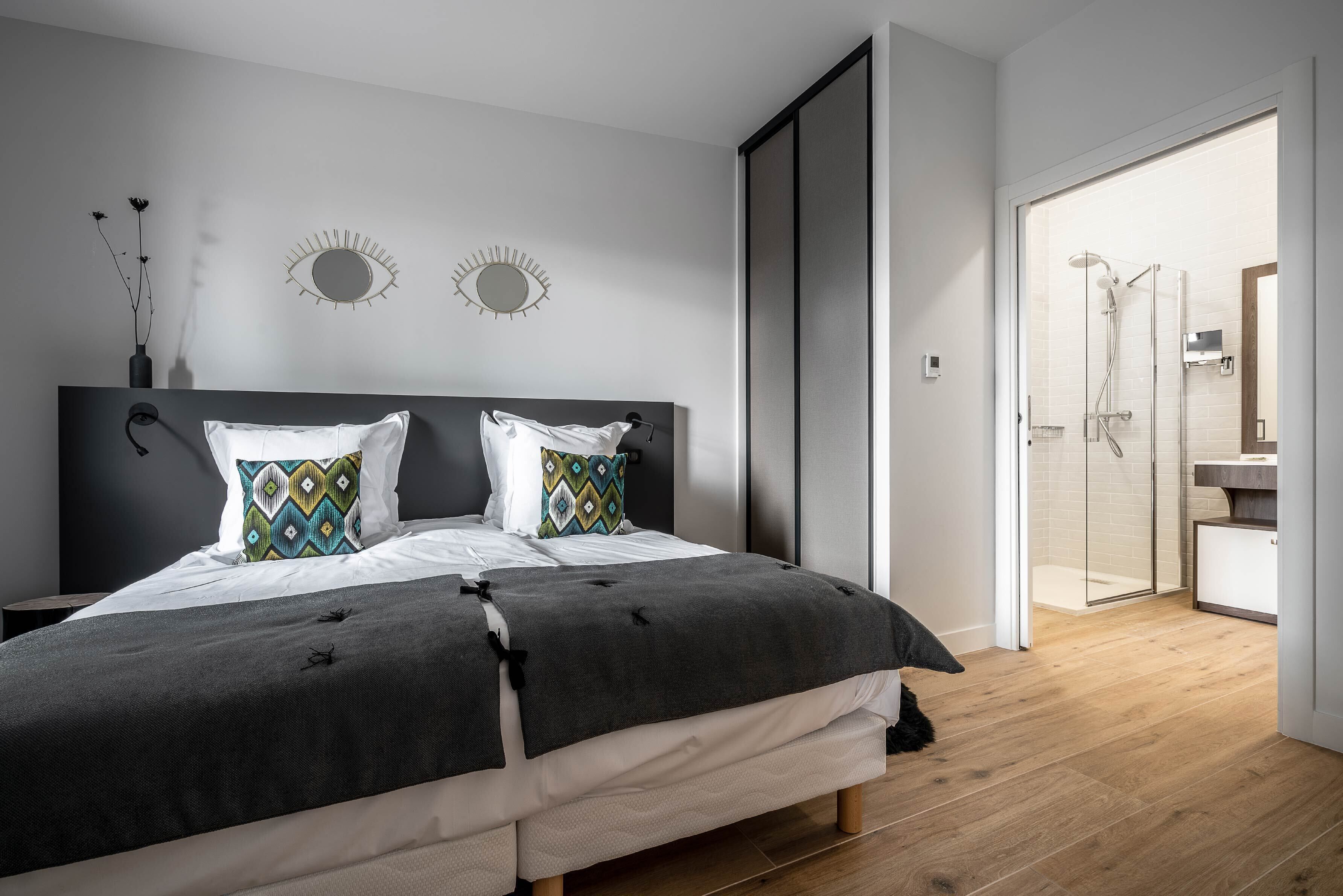 A modern bedroom with a large bed, two decorative wall mirrors, and a view into a bathroom with a glass shower.