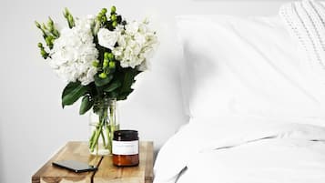 A wooden nightstand with a smartphone, a jar of candles, and a vase of white flowers against a white wall.