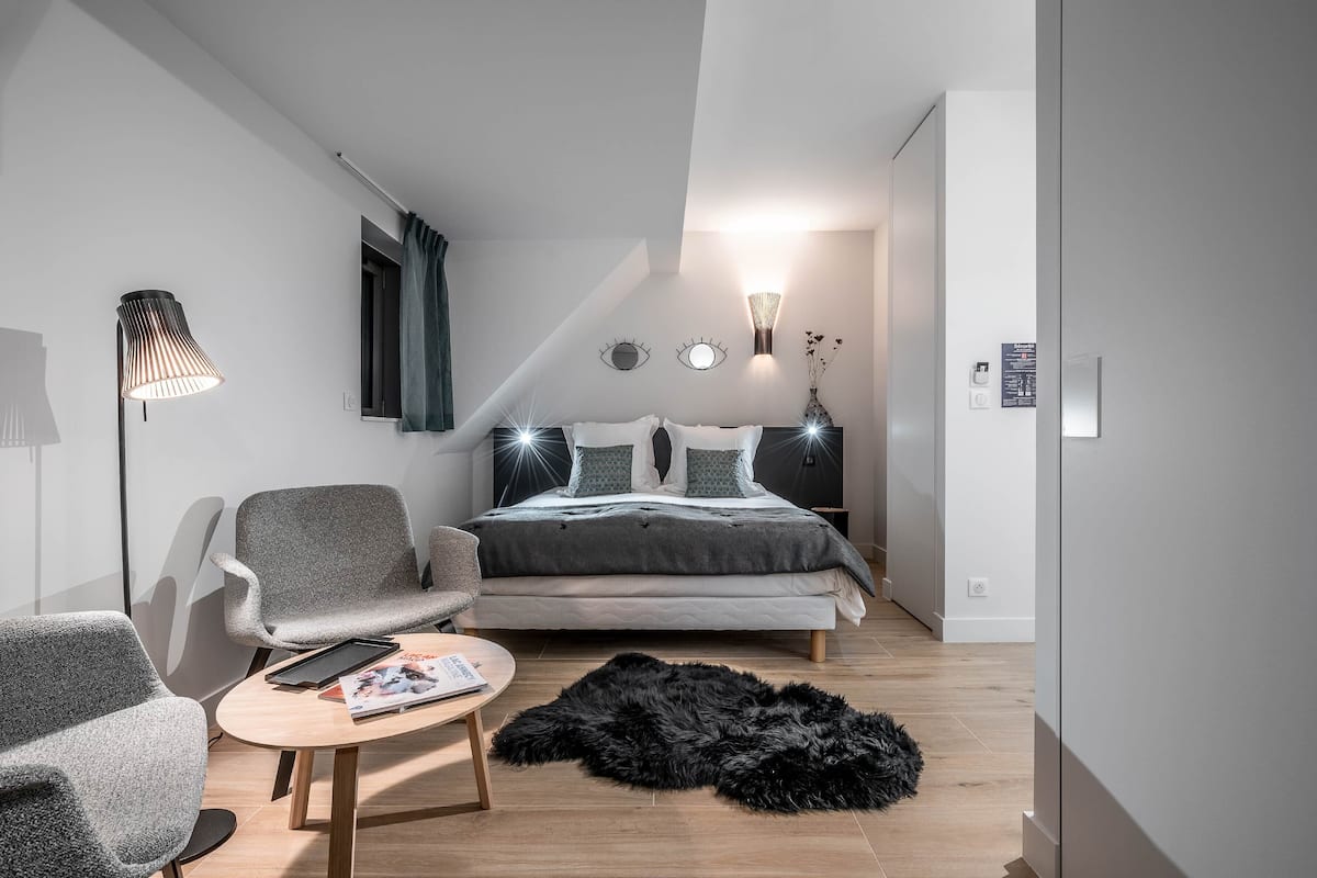 Une chambre moderne avec un lit, deux fauteuils gris, une petite table ronde, un tapis noir à poils longs et une lampe fixée au mur.