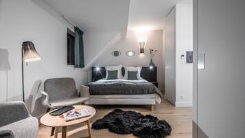 Ein modernes Schlafzimmer mit einem Bett, zwei grauen Sesseln, einem kleinen runden Tisch, einem schwarzen, flauschigen Teppich und einer Wandlampe.