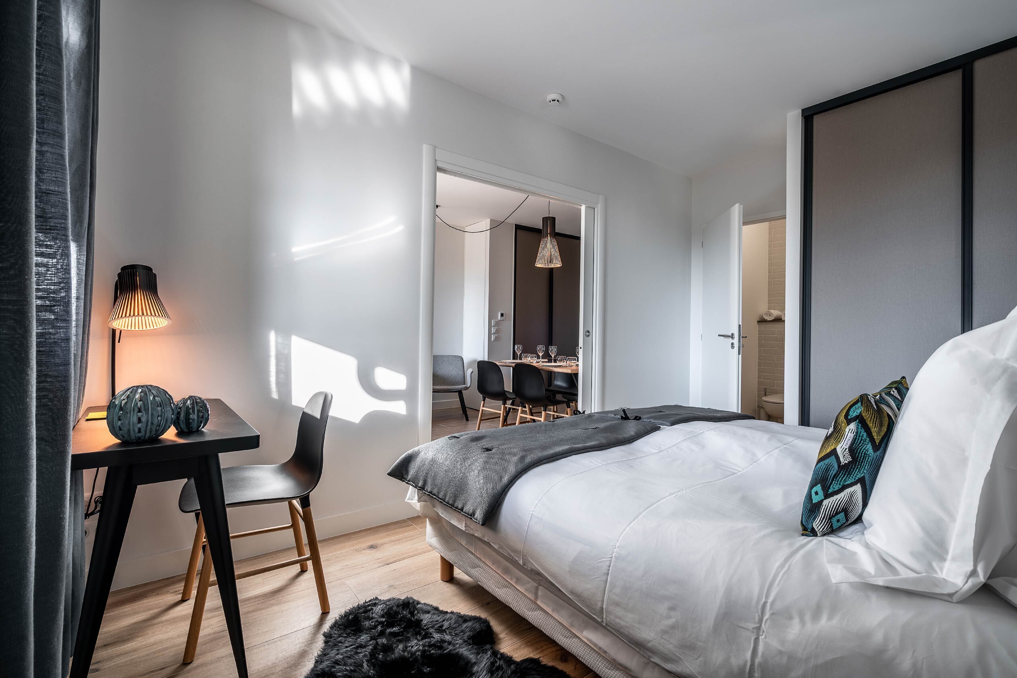 Quarto moderno com cama grande, escrivaninha com luminária e área de refeições ao fundo.