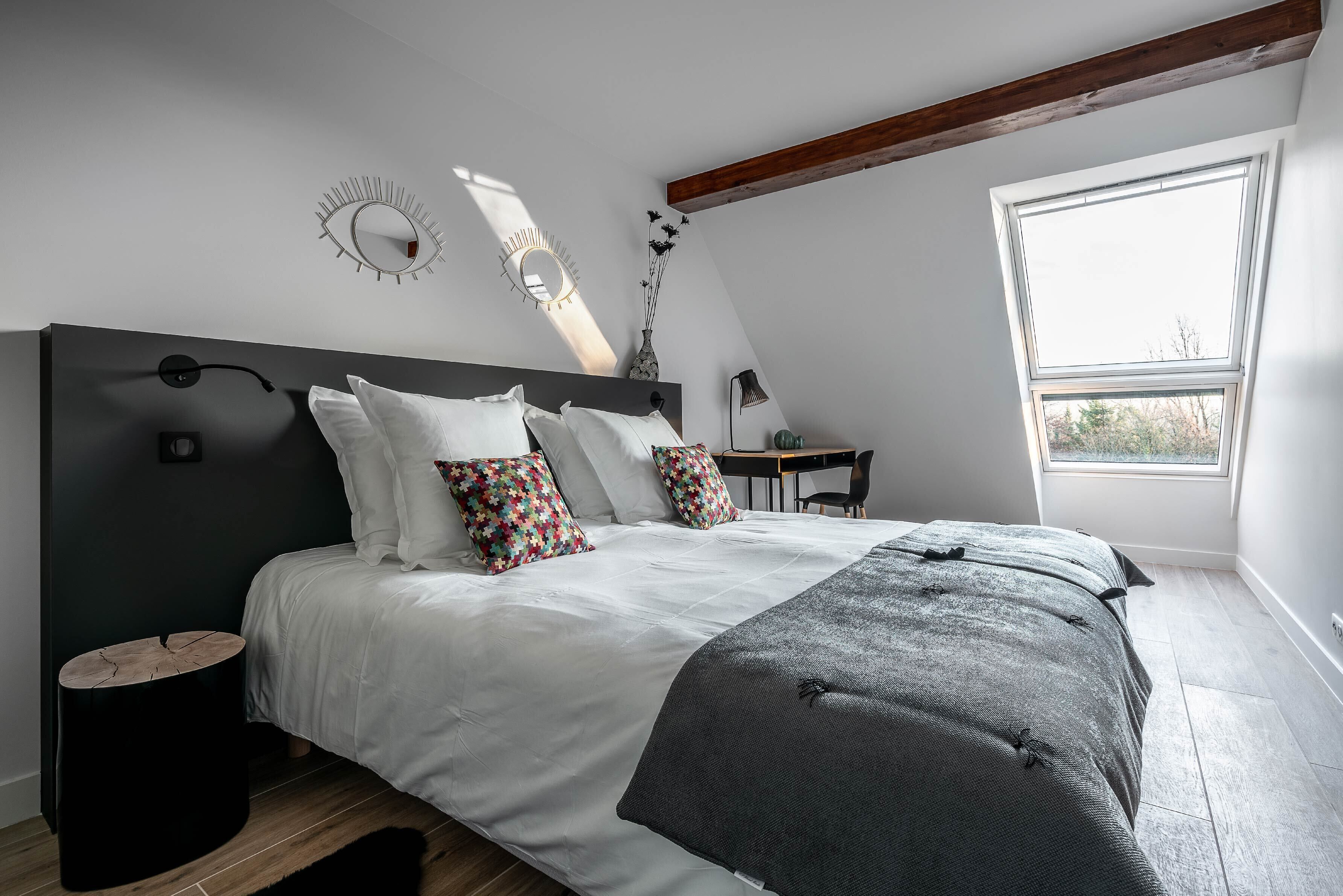 Une chambre à coucher avec un grand lit, une tête de lit en bois, une petite table de chevet et une fenêtre donnant sur des arbres.