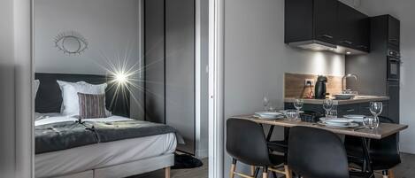 Une chambre moderne avec un lit, une table de chevet et une commode. Un coin repas avec une table, des chaises, et un espace cuisine avec des placards et des appareils électroménagers.