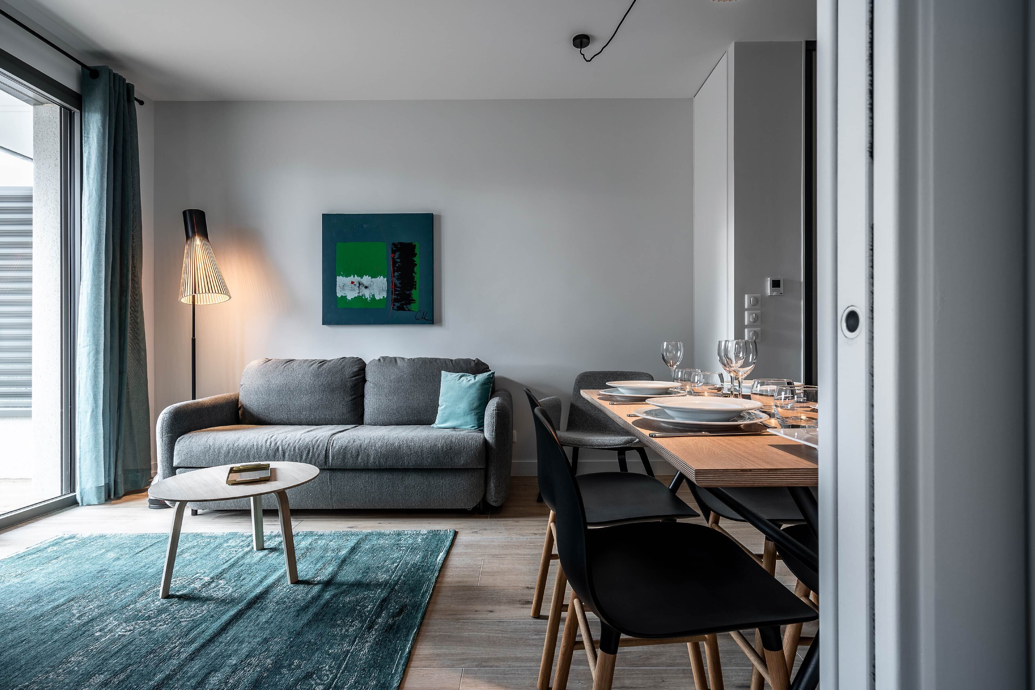 A modern living room with a grey sofa, a small round table, and a dining area with a wooden table and chairs.