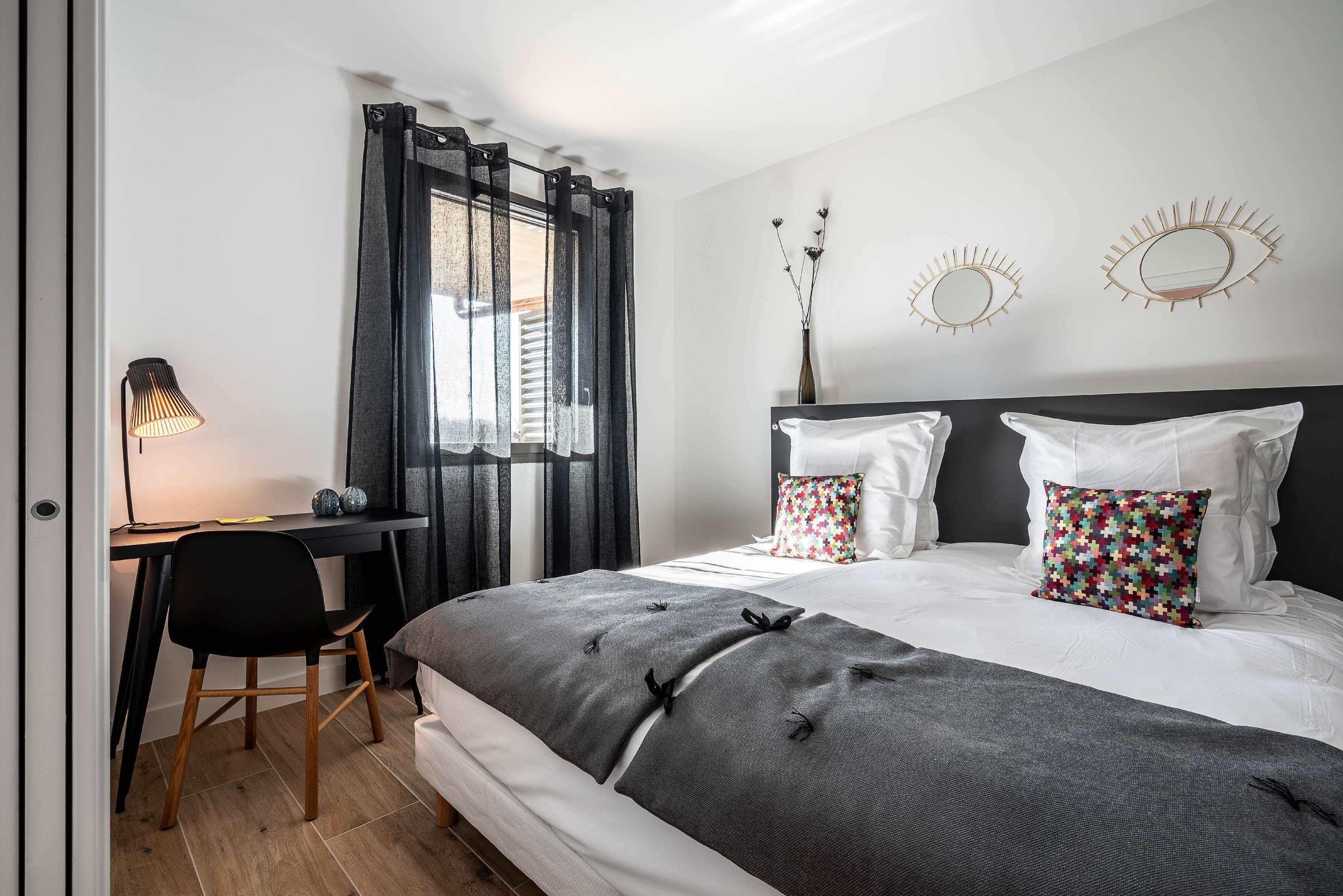 A bedroom with a bed, a desk, and a chair. There are curtains on the window, and a lamp on the desk.