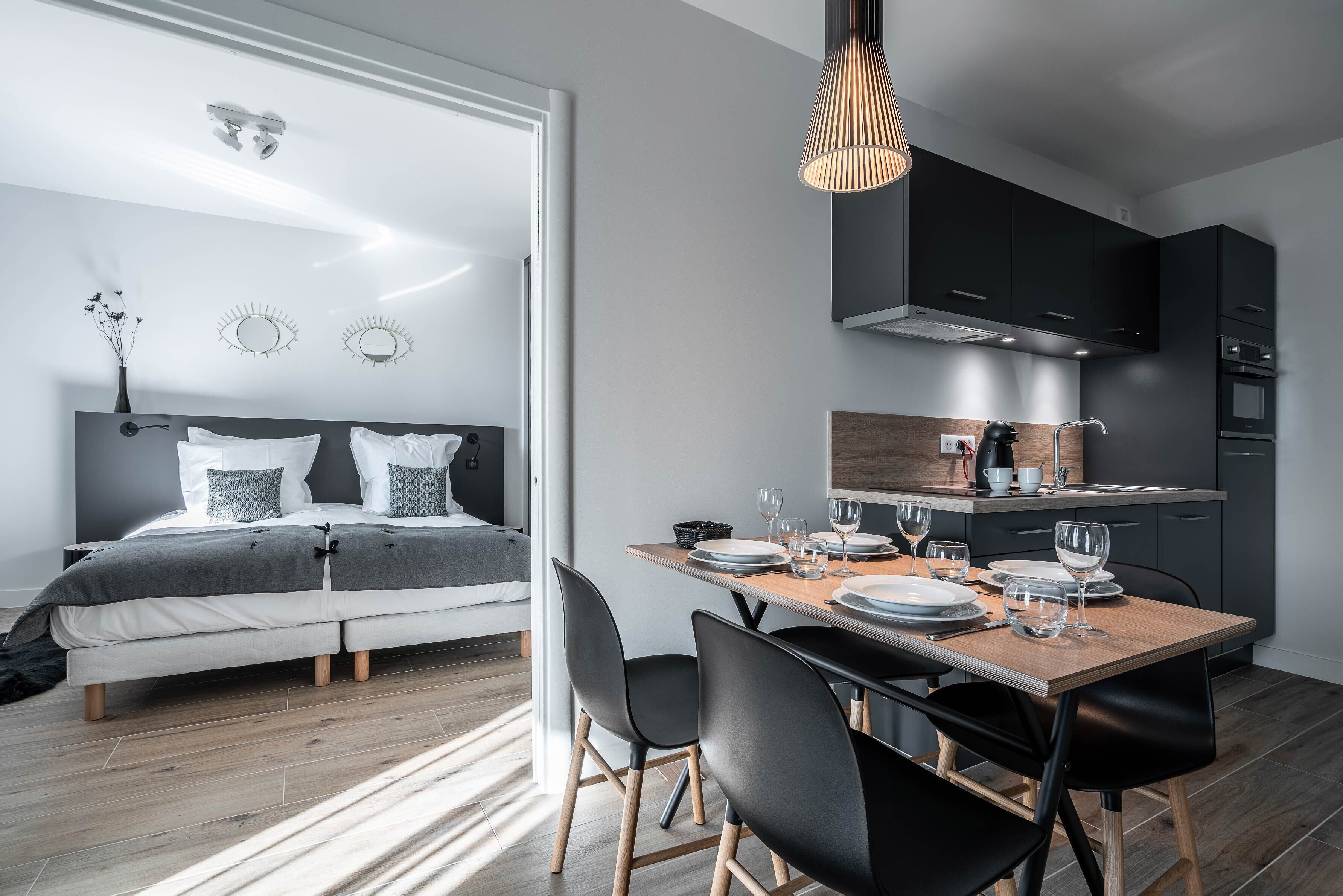 A modern bedroom with a large bed, bedside tables, and a nightstand. There is a dining area with a wooden table, chairs, and a kitchen with dark cabinets and appliances.