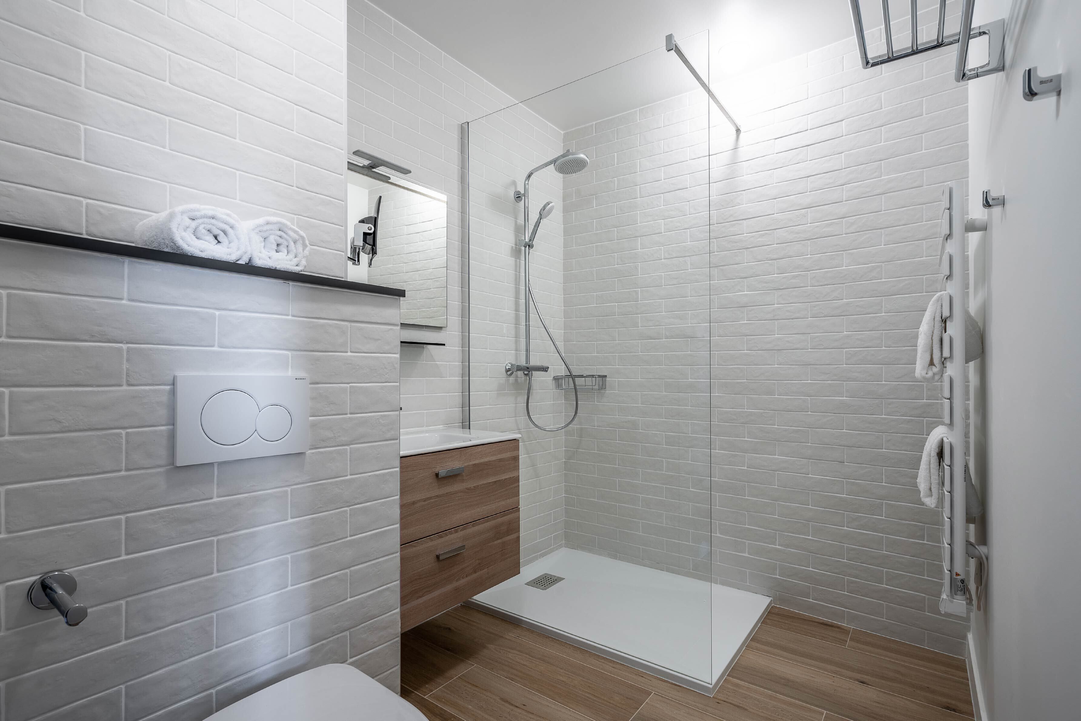 A modern bathroom with white brick walls, a glass shower enclosure, a wooden vanity, and a towel rack.