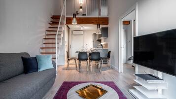 A modern living room with a grey sofa, a dining table with chairs, a staircase, and a flat-screen TV.