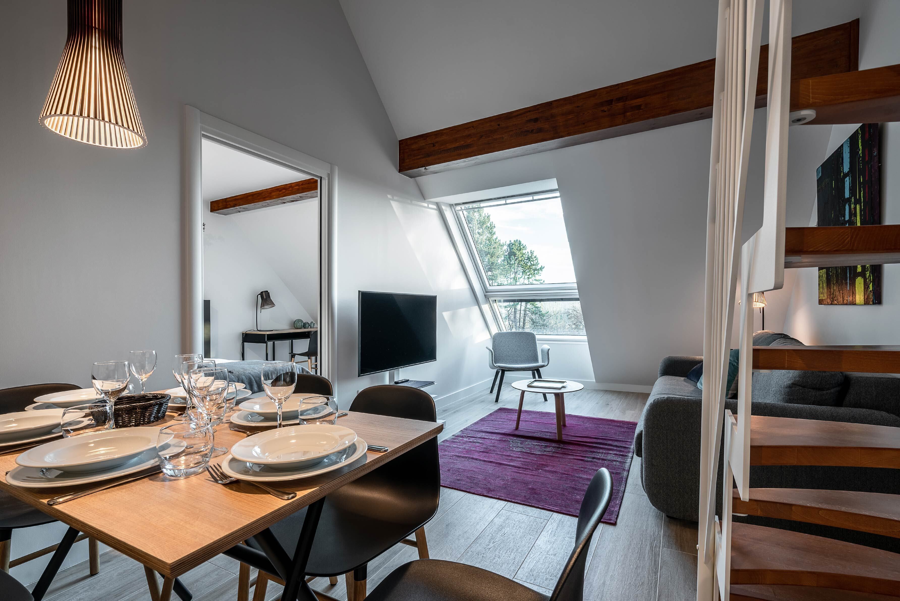 Een moderne eethoek met een houten tafel voor zes personen, een televisie en een comfortabele zithoek met een bank en fauteuils.